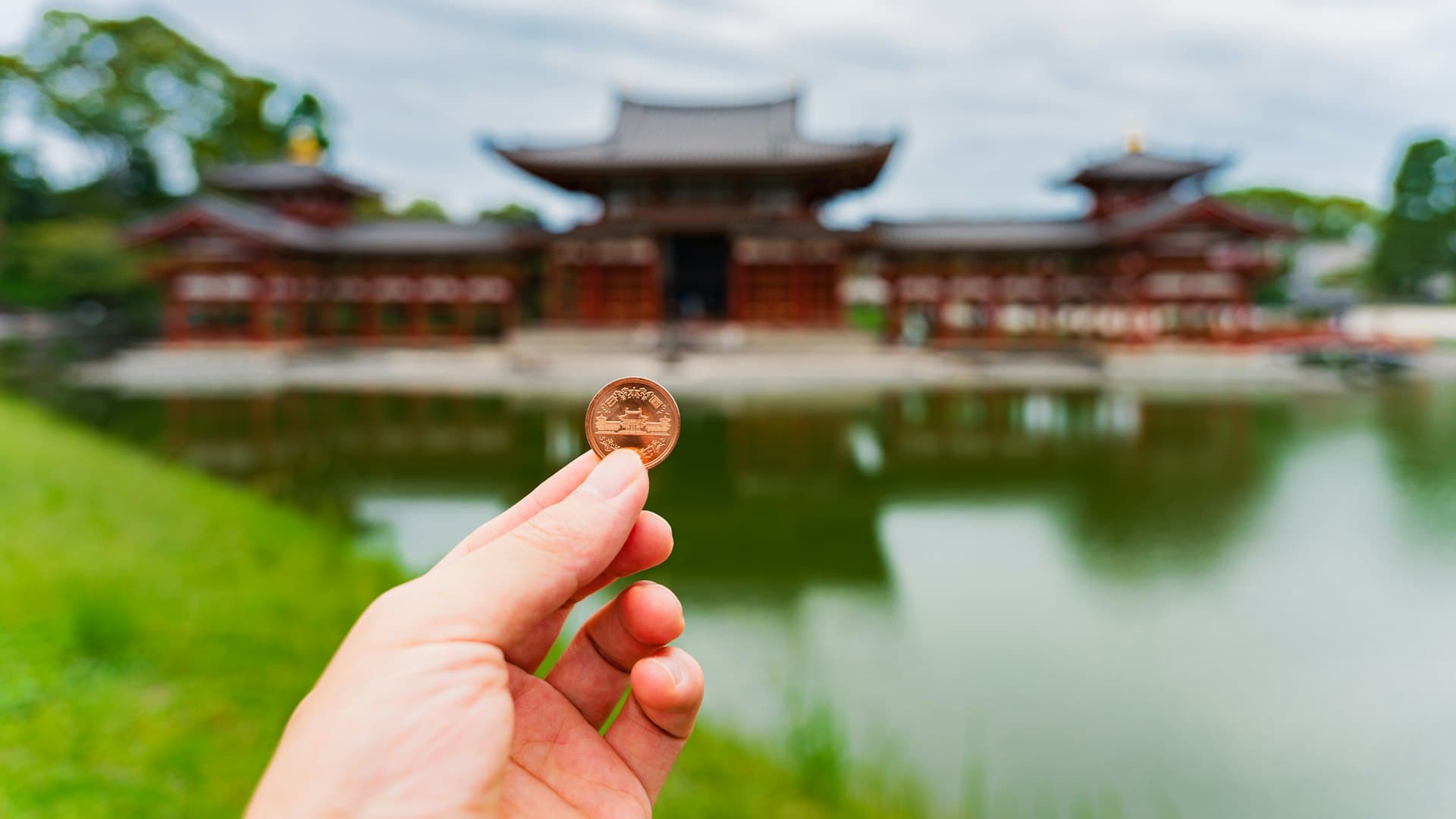 Hand holding a Japanese yen coin in front of Byodo-in temple